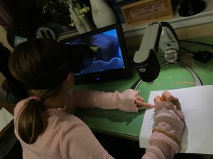 A girl leans close viewing a monitor that shows her hand on the left of frame. To the right of the frame her hand holds a pencil over a worksheet. The worksheet is underneath the document camera of a CCTV system.